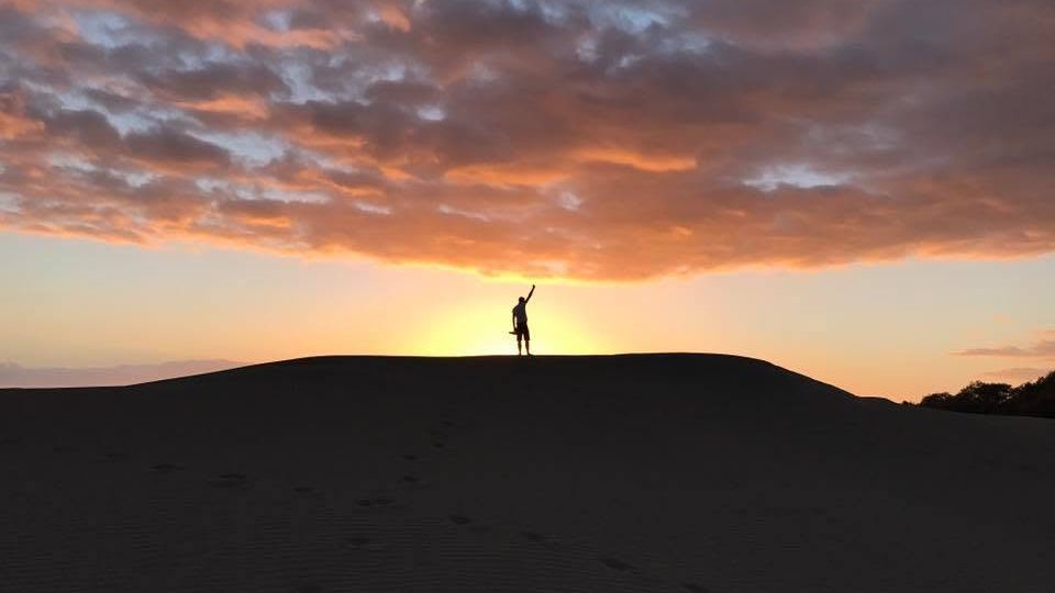 Silhouette d'un voyageur seul au sommet d'une dune, bras levé face au coucher de soleil aux Canaries